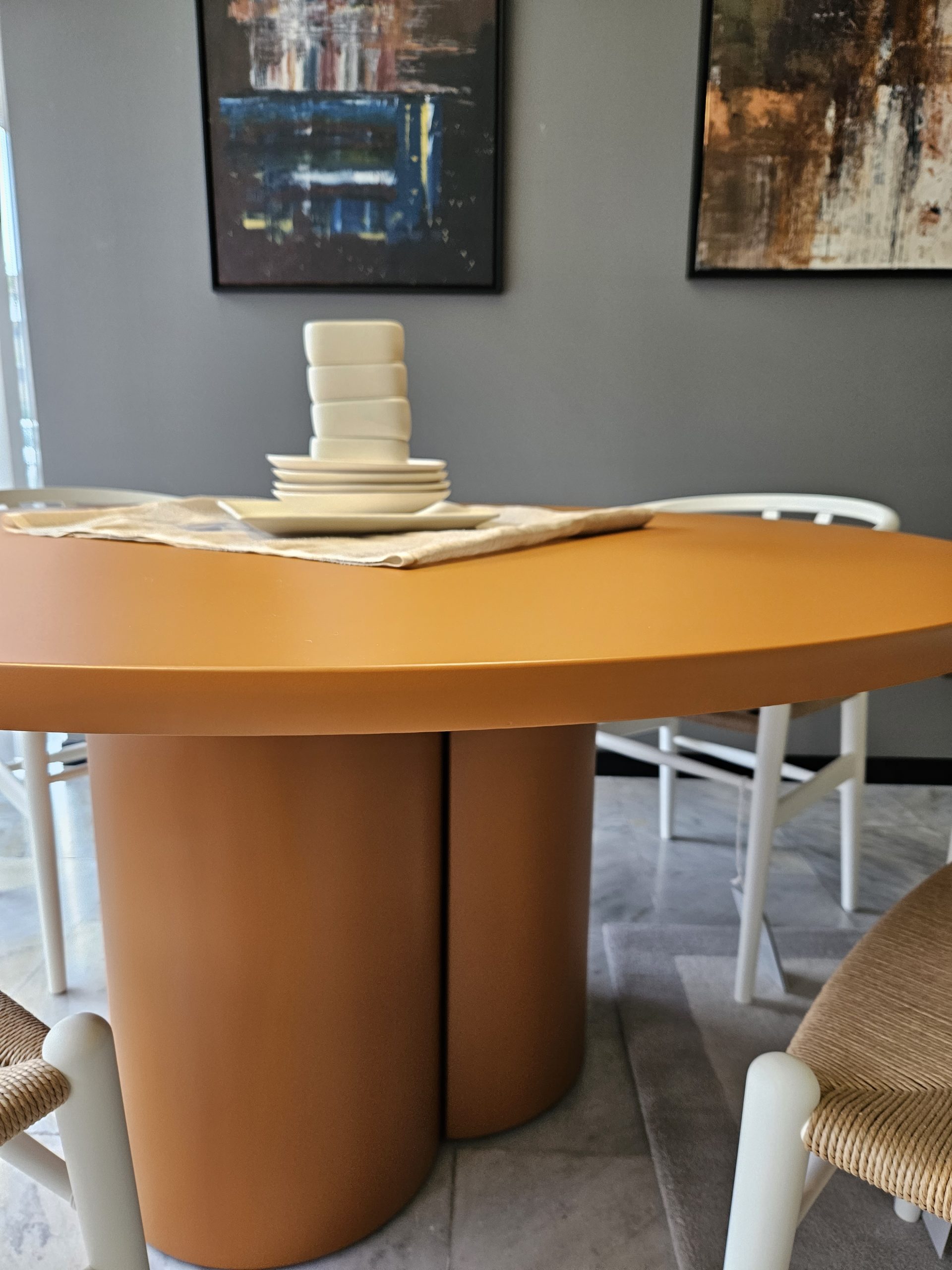 Ein modernes Esszimmer mit einem runden, orangefarbenen Tisch mit schlankem, zylindrischem Untergestell. Weiße Stühle mit geflochtenen Sitzflächen umgeben den Tisch. Teller und Schüsseln in neutralen Farben stapeln sich auf einem beigen Tischläufer. Abstrakte Kunstwerke schmücken die grauen Wände.