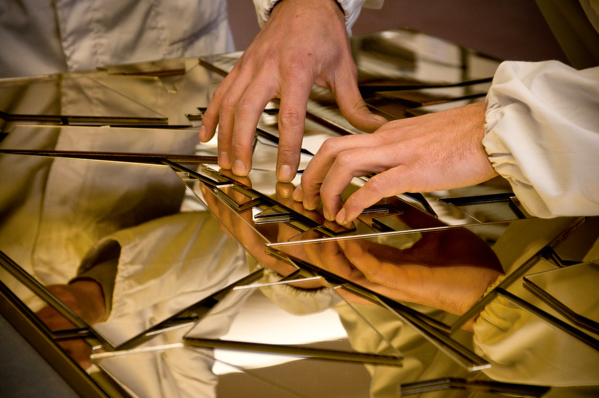 Two individuals in lab coats gently touch a golden, reflective surface with geometric patterns. Their hands and the intricate design are mirrored in the surface, creating an abstract and visually striking composition.