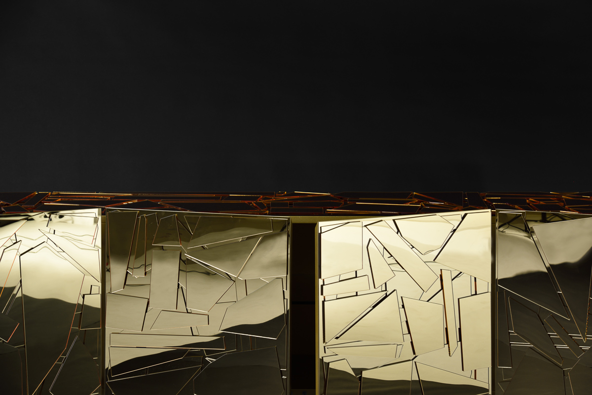Close-up of a gold-colored, geometric-patterned surface against a dark background. The surface has an abstract, fragmented design, reflecting light and casting shadows, creating a dynamic visual texture.