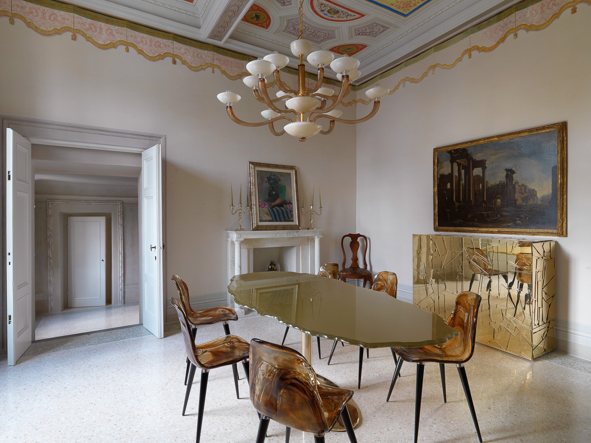 Elegant dining room with an oval glass table, seven modern chairs, and a large chandelier. Two paintings adorn the walls, with a gold-toned cabinet beneath one. An open doorway and ornate ceiling add to the refined atmosphere.
