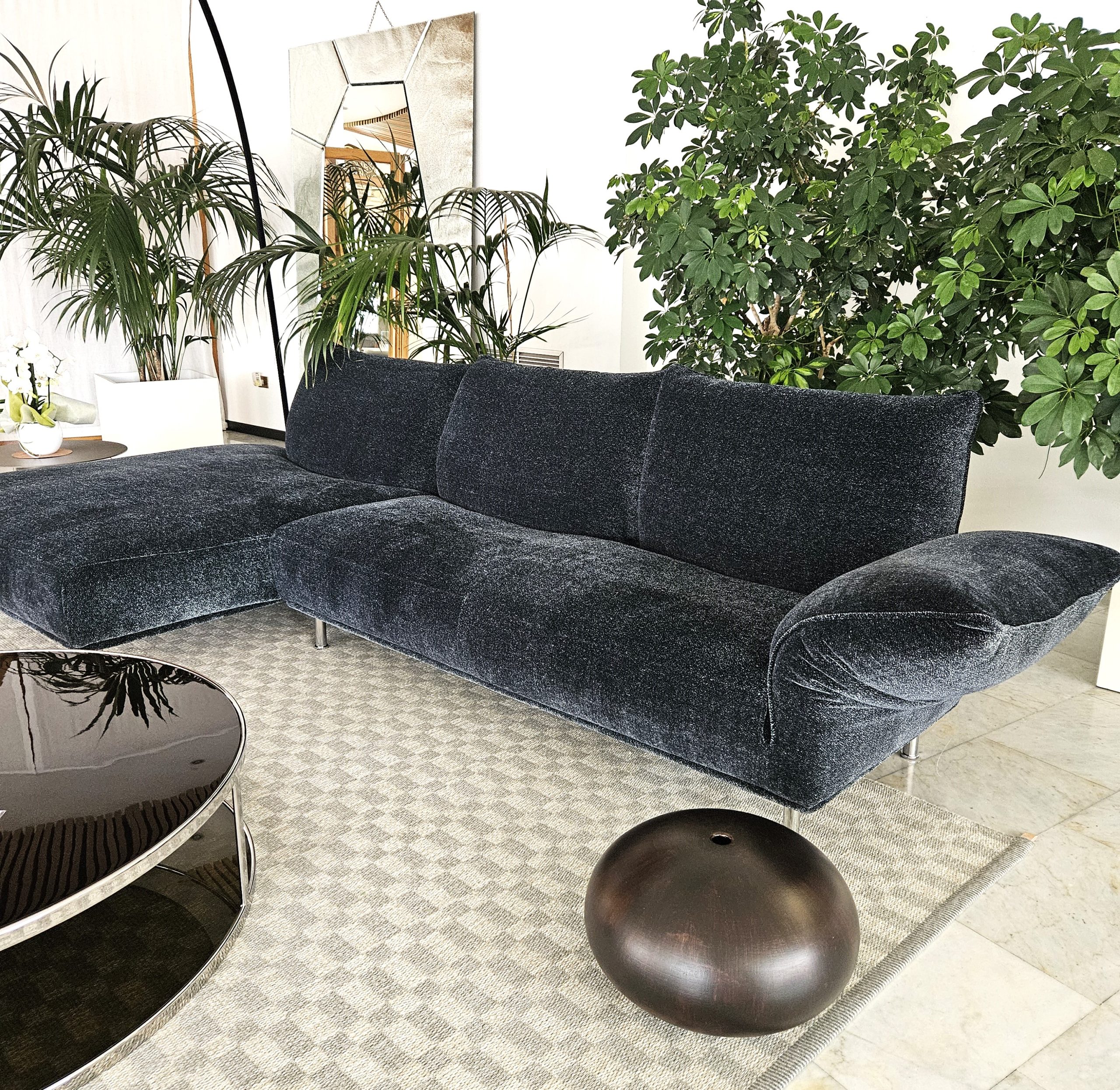 A modern living room featuring a dark blue velvet sofa on a checkered light gray rug. A round, glossy black coffee table and a black spherical object sit nearby. Lush green plants and a geometric mirror decorate the white walls.