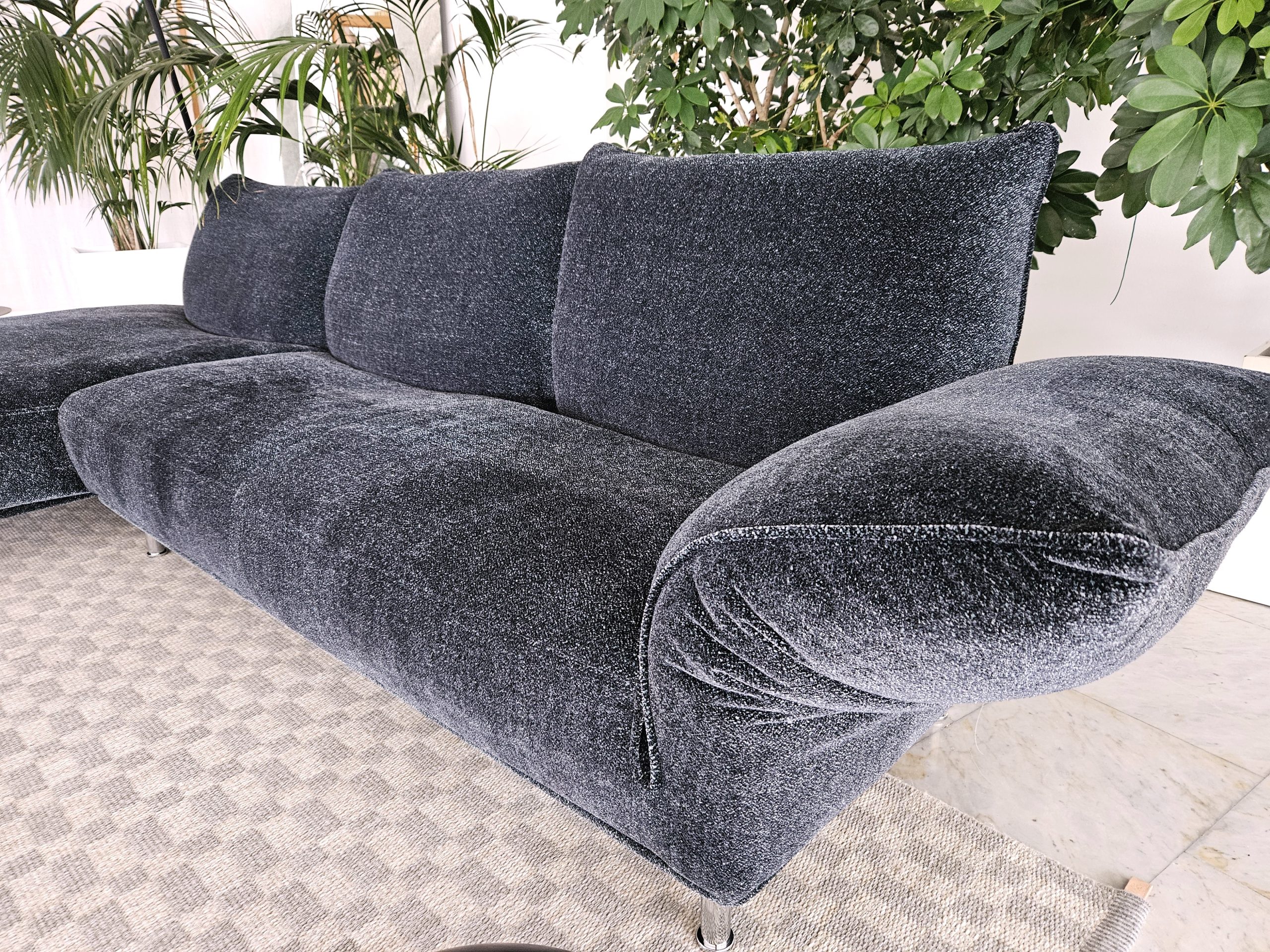 A modern, plush, dark gray sofa with curved armrests and a textured fabric sits on a checkered patterned rug. Lush green plants are visible in the background, adding a touch of nature to the room.