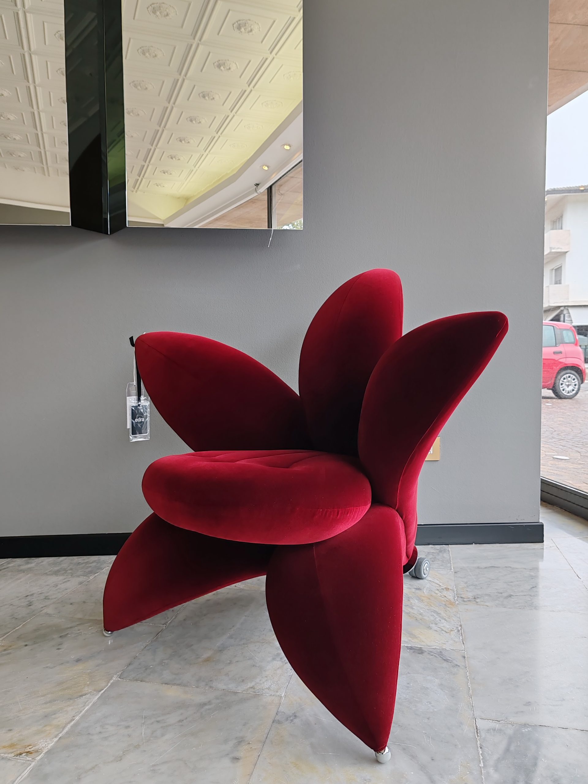 Ein moderner Stuhl mit tiefroten, blütenförmigen Kissen, die wie eine Blume angeordnet sind, steht in einem minimalistischen Raum mit grauen Wänden, Marmorböden und einem großen Fenster mit Blick auf ein geparktes rotes Auto draußen.